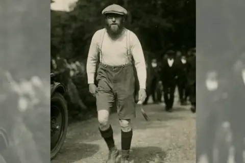 Manx National Heritage An old portrait photograph shows a man walking wearing dungaree shorts, with socks up to his knees and old walking boots. He is also wearing a flat cap, with a bushy beard and is looking at the camera.