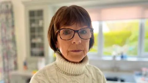 A woman with shoulder length brown hair looks at the camera, wearing brown rimmed, clear glasses. She wears a cream turtleneck. Behind her is a out of focus kitchen sink, with a window above it. A green hedge can be seen outside.