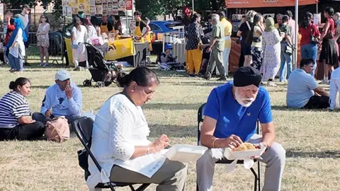 Art Asia Crowds of people gather around food stalls on a field in a music festival. In the foreground, a man and a woman are eating out of takeaway boxes. 