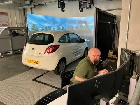 A man sat a a bank of computers with a car in front of big screens in the background