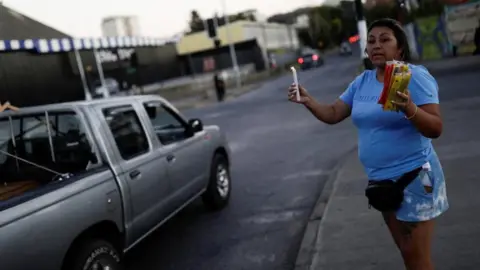 Reuters A woman wearing a light blue T-shirt and matching shorts stands on a pavement in Concepción, Chile, holding a lit candle in one hand and packs of candles for sale in the other hand. A silver pick-up truck is cruising past.