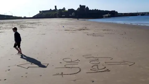 A silhouette of a child walking away from Manx writing in the sand with two arrows drawn on either side, you can see Peel Castle in the background.