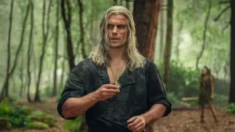 Netflix Henry Cavill como Geralt com longos cabelos grisalhos e cortes em todo o rosto. Ele segura um pingente de prata. Ele está vestindo uma camisa verde de botões abertos e mangas arregaçadas. Ele está em uma floresta.