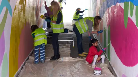 A number of people painting an underpass, with white walls and colour on them. They are all bending over, wearing covers and holding paint brushes. A cover is on the floor. 