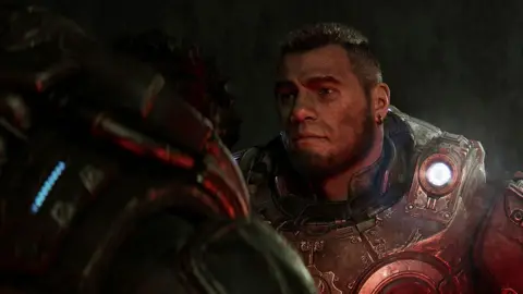 Microsoft CGI shot from Gears of War: E-Day shows a man in heavy, space-marine armour listening intently to another character, whose shoulder the viewer is looking over. It's a dark scene, lit only by a torch built in to the shoulder of the main subject's armour.