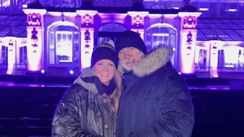 Paul Carlier A husband and wife stand wrapped up in winter clothes in front of an illuminated building. 