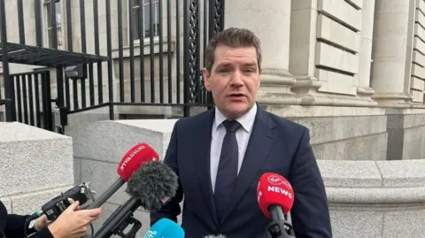 PA Media Peter Burke, a man with fair hair, wearing a dark suit, dark tie and grey shirt. There are microphones in front of him and grey buildings in the background.