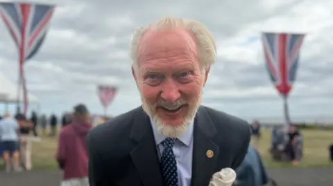 BBC/Jim Scott Brian Burnie, who has fair blonde hair and a beard and is wearing a blue blazer on top of a blue-striped shirt and patterned tie, originally commissioned the statue as a tribute to his friend, Len Gibson. He is standing on the path in front of the field where a service has been held. In the background are three large poles carrying the Union Jack.