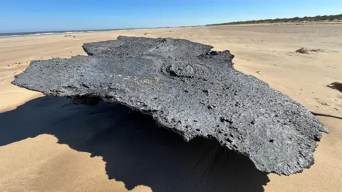 A large clump of charred plastic sat on a golden sandy beach. The sky is blue. Sand dunes are to the right of the plastic and the sea to the left.