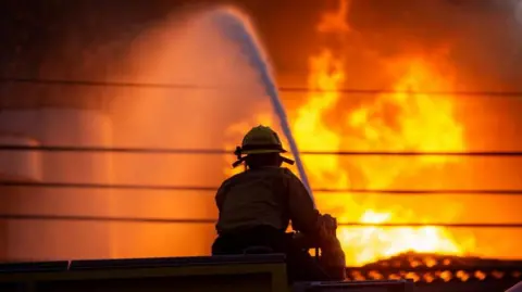 A firefighter tackling a fire in the Palisades area of Los Angeles. They sit in the foreground with their back to the camera holding a hose. A stream of water is going from the hose towards a tire in the roof of a building in the background. 