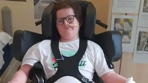 Flemen family Daniel Flemen in a hospital room strapped into a wheelchair with head and shoulder supports. He has dark hair and glasses and a tube in his nose.