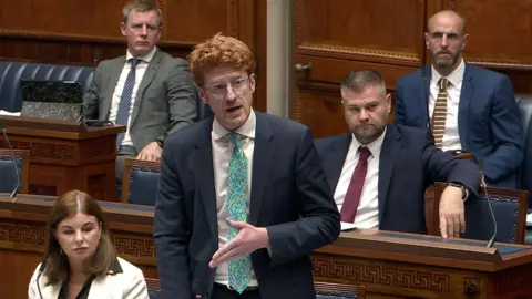 Matthew O'Toole with ginger hair, standing in the Assembly chamber wearing round spectacles and a navy suit with green tie