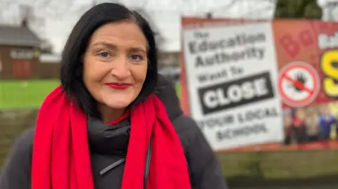 Les is smiling into the camera. She has bobbed black hair. She's wearing red lipstick and a red scarf. Her black coat is padded. A sign opposing the closure of the school is out of focus behind her.