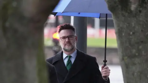 PA Media TV auctioneer Charles Hanson wearing a black coat holding a umbrella 