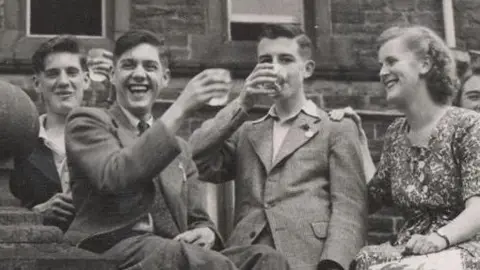 University of Lancashire A black and white photo of three men and woman holding up glasses while sat on a wall outdoors. 