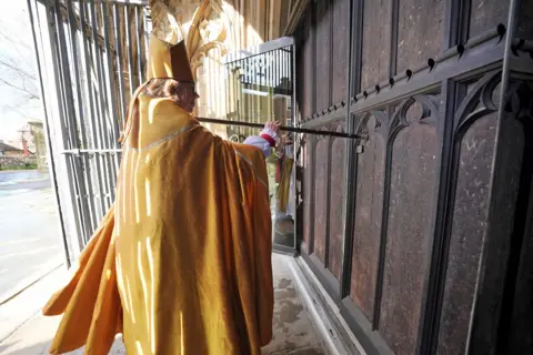 Yui Mok / PA Media Dame Sarah in robes strikes large wooden door of cathedral with staff as sunlight pours over the scene