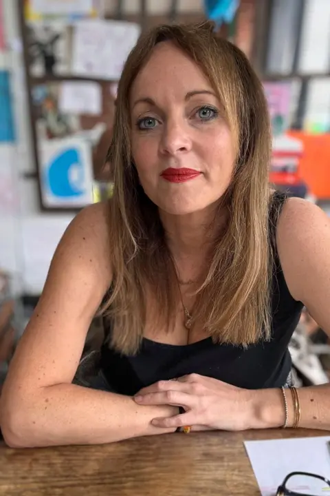 Jenny Evans Jenny Evans looks at the camera, she is wearing red lipstick and has straight blonde hair. She is sat at a table resting her crossed arms on the table. 