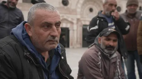 BBC Two men sit next to each other in a square in Jordan with several men standing behind them