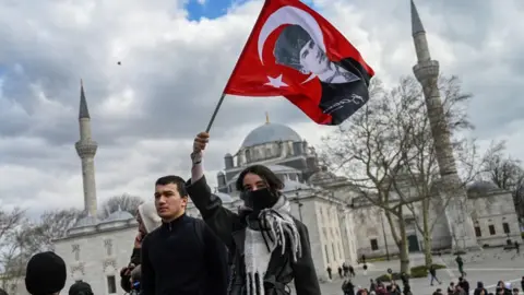 GETTY IMAGE Seorang wanita mengawasi bendera poster Mustafa Kemal Ataturk sebagai mahasiswa universitas menunjukkan menentang keputusan Diploma Metropolitan Metropolitan Istanbul pada 19 Maret 2025 di Gerbang Utama Universitas Istanbul di Istanbul