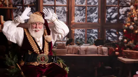 Danielle Axworthy Allan Evans dressed as Santa, sticking his tongue out with his hands in the air, in front of a snowy window background and a Christmas tree