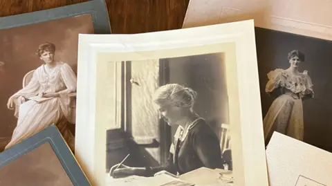 Jebb Family A collection of black-and-white photographs of Eglantyne Jebb. The main photo shows her sitting at a desk and writing. Other photographs show her as a young woman wearing long formal dresses. The photos are laid out on a brown wooden desk. 
