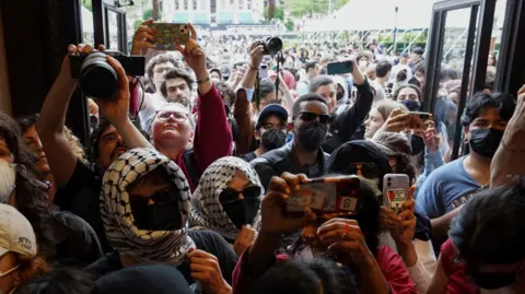 Protesters, some wearing face masks, sunglasses and head scarfs, enter an open door, with dozens more protesters behind them.