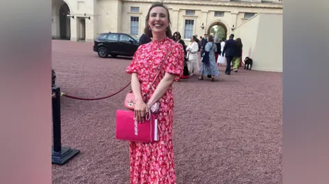 A smiling Rebecca inside the grounds of Buckingham Palace. She is wearing a pink flowers dress. She is holding a pink folder from the Lorraine show.