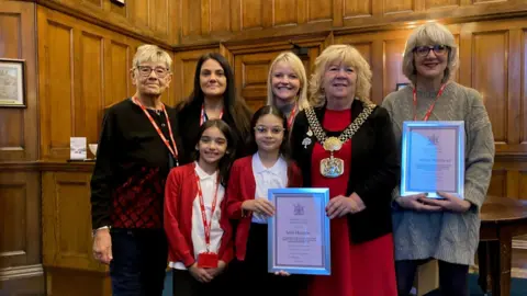 Two children and five adults holding two framed bravery certificates in a wooden-panelled room. 