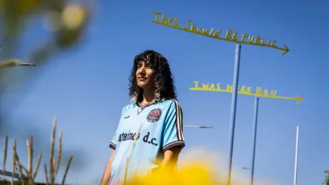 Turner Prize-winning artist Jasleen Kaur photographed on the banks of the River Clyde next to two weather vein sculptures which form part of her new "Superstructure" commission. She is standing in front of the weather veins and has long dark, curly hair. She is weather a Fontaines DC football top. The weather veins are metal arrows with text on them. One reads "This should be public" and the other says "I was here b4 you".