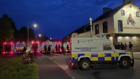 A line of police in riot gear. It's night. A number of police land rovers are parked. 