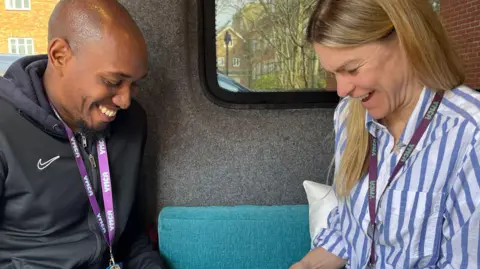 BBC/Julia Gregory Marlon Williams (left, a man in a black tracksuit with a purple lanyard) and Fiona Oldam (right, a woman in a blue and white striped top with a purple lanyard) both smiling and looking down 