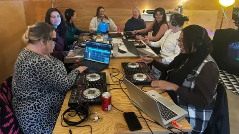 Cad Taylor/ BBC A group of women are round a table with lots of media and DJ equipment.