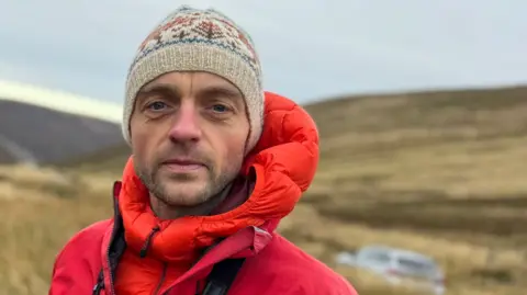 Alex Priestley, who has a short, stubbly beard. is wearing a red hooded puffer jacket underneath a red windcheater. He has a brown woollen hat on his head. He is standing in the hills under a grey sky. There is no snow.