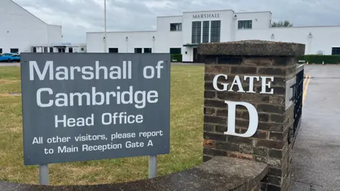 A large grey sign in the foreground says "Marshall of Cambridge Head Office". It is next to a brick pillar with "Gate D" on it. In the background is the company's white Art Deco reception building.