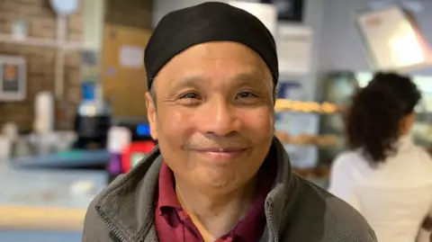 A man in a grey jacket and black hat smiles at the camera with the cafe blurred in the background. 
