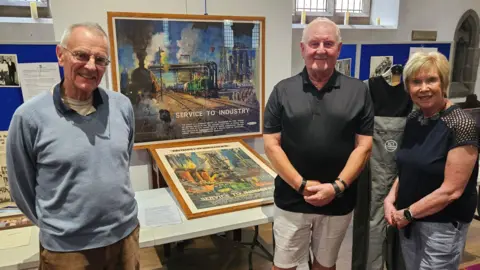 Three people; to the left is an elderly man with receding grey hair. He is wearing a pale blue jumper or polo shirt over a white t-shirt. To his right is an elderly man, also with receding grey hair and wearing a short-sleeved black shirt and white shorts. On the far right is a woman with short blonde hair, wearing a black top and light blue shorts or trousers. All three are smiling. Behind them are stands with old photographs and documents, and in a prominent position between them are two colourful old railway posters.