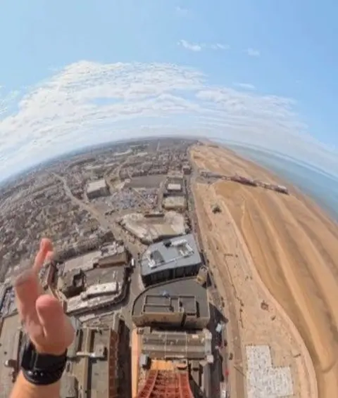 LMSD (Let’s Make Stupid Decisions) A view of Blackpool and the beach from the top of the tower, taken from a camera worn by one of the men as he jumps. His hand can be seen in the foreground.