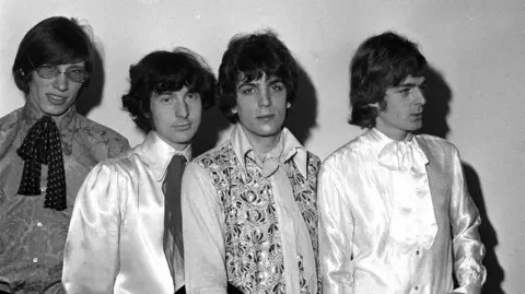 Pink Floyd's Roger Waters, Nick Mason, Syd Barrett and Rick Wright stood in a line wearing satin shirts and neck ties. The photograph is black and white.