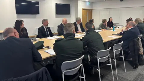 A meeting room with a long wooden table. There are men and women in suits and jackets sitting round the table. There are two TVs on the wall.