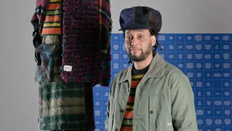 Julie Howden Nicholas Daley, wearing a khaki jacket and colourful striped knitted jumper, and a denim hat, stands beside a mannequin dressed in wool plaid and knitted garments in autumnal colours. There's a blue patterned background behind him.