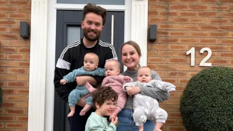 handout Jamie and Hayley pose outside a house holding their babies with son Otis standing in front smiling and holding his baby sister's foot.