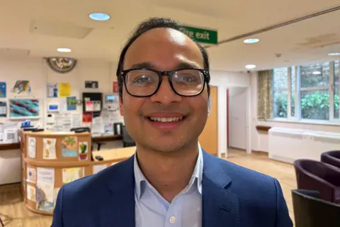 Man wearing black rimmed glasses, blue blazer and light blue shirt, black short hair, smiling at the camera. In the background is a reception desk, window, and back wall with pictures and posters on it.