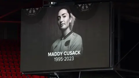 Getty Images A screen displays a tribute in memory of former Sheffield United player Maddy Cusack during a match between Sheffield United and Crystal Palace. It's a black and white photo of Maddy in her kit, her hair tied back in a pony tail. Beneath it reads: Maddy Cusack 1995-2023.