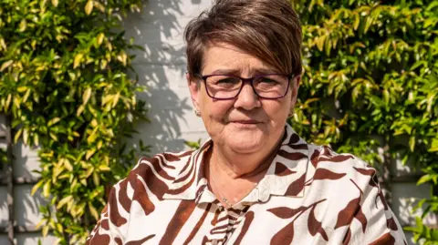 Bhasker Solanki A head and shoulders picture of Tracy Page, wearing a brown and cream patterned top, sitting in front of a wall with climbing plants.