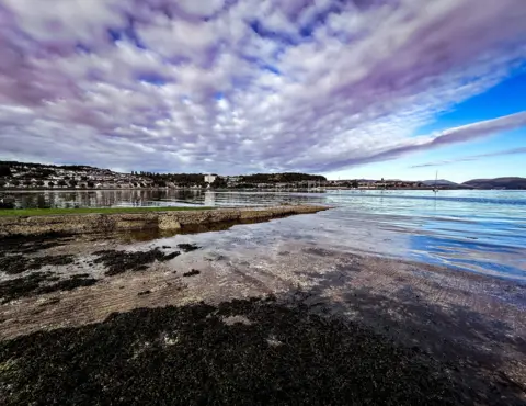 Tommy Murphy Bamford Panoramic image of white clouds over a bay, reflected in the water with houses in the far distance