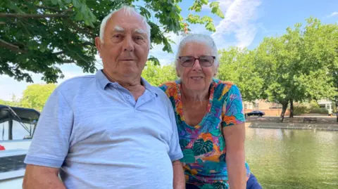 Alex Pope/BBC David and Joy Nottingham, siting on a boat on a tree-lined river, smiling and looking at the camera. David is wearing a blue polo shirt and Joy is wearing a bright, colourful floral top. 