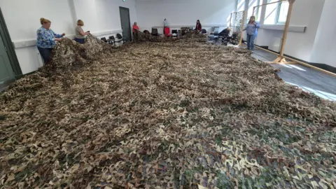 A huge green and tan camouflage net, covering the entire floor of a room. Women stand at each end of the net, lifting parts of it up off the floor. 
