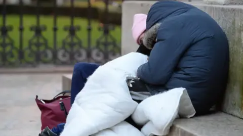 PA Media Two people are rough sleeping on some steps. One is wearing a blue jacket, the other a pink hat. 