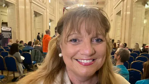 A woman with fair hair and a light coat smiles at the camera, behind her is a room full of people