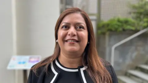 Deputy Munazza Malik, a lady with brown hair wearing a black jacket and dress and white pearl earrings.
